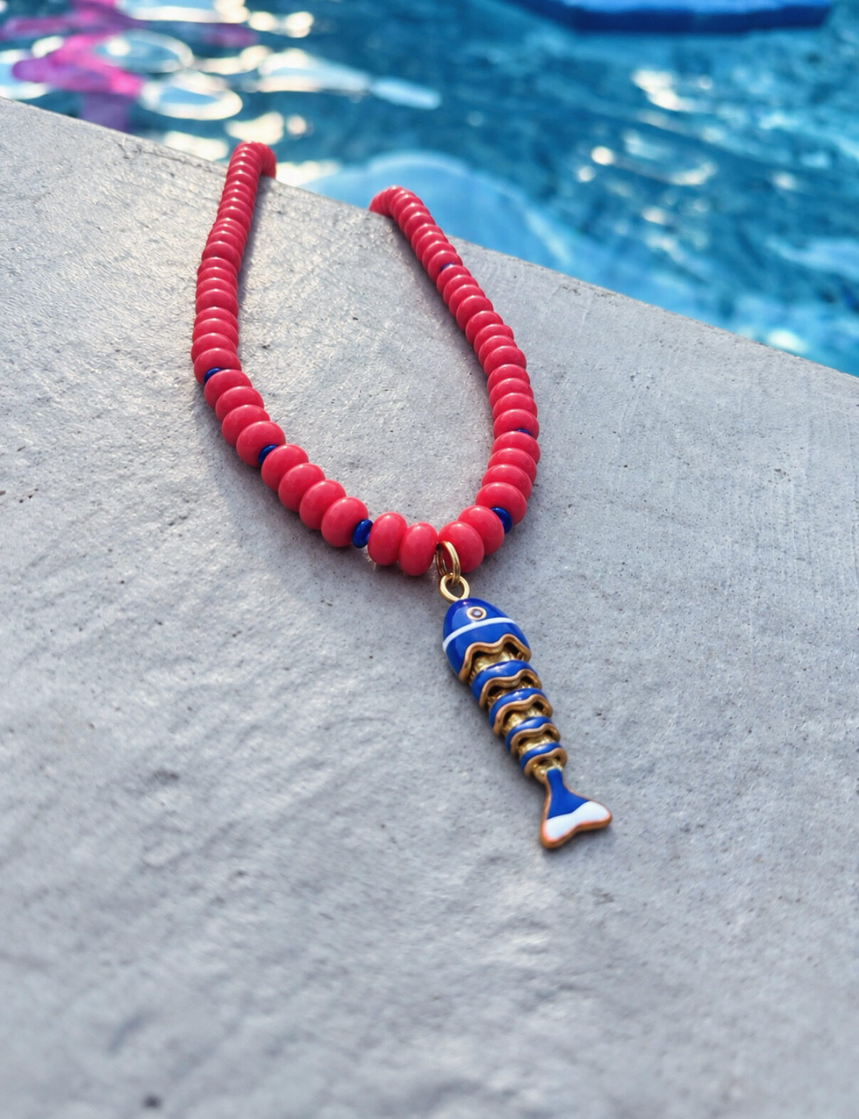 Coral beaded necklace with a blue enamel fish pendant beside a pool