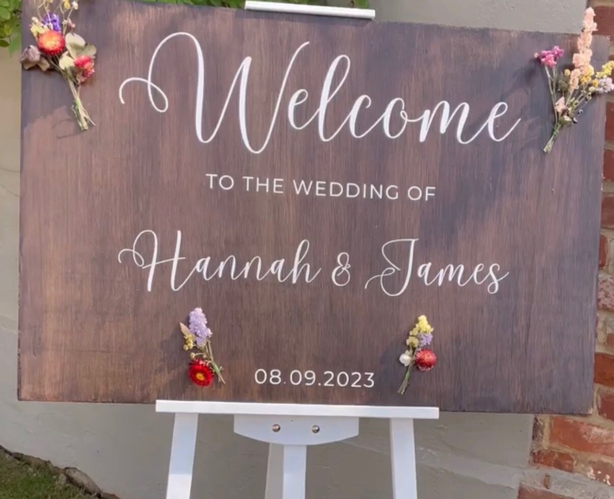 Stained wood wedding welcome sign for Hannah and James with white script and dried flowers on a white easel