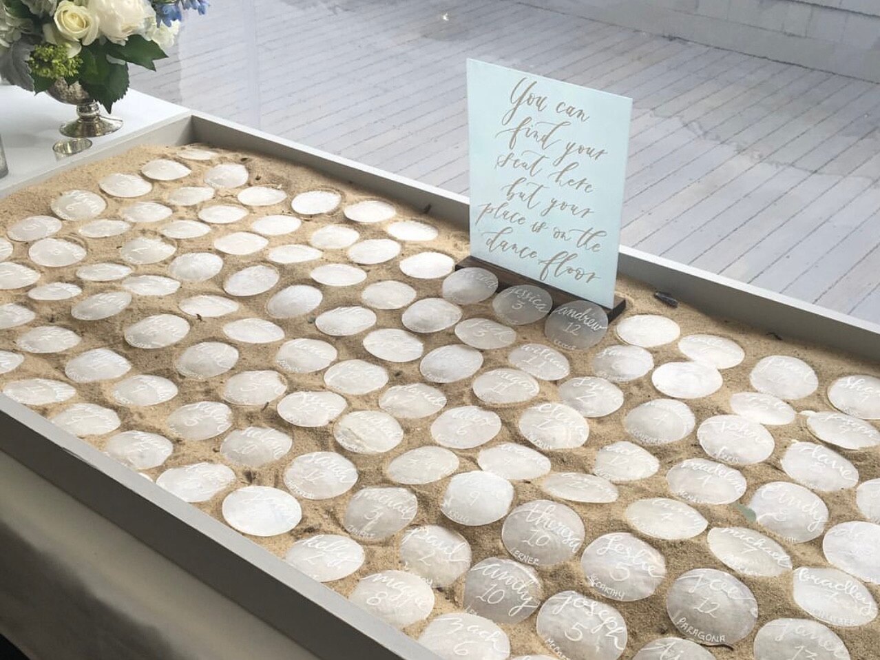 Hand-lettered capiz shell escort cards set in a tray of sand with a find your seat sign